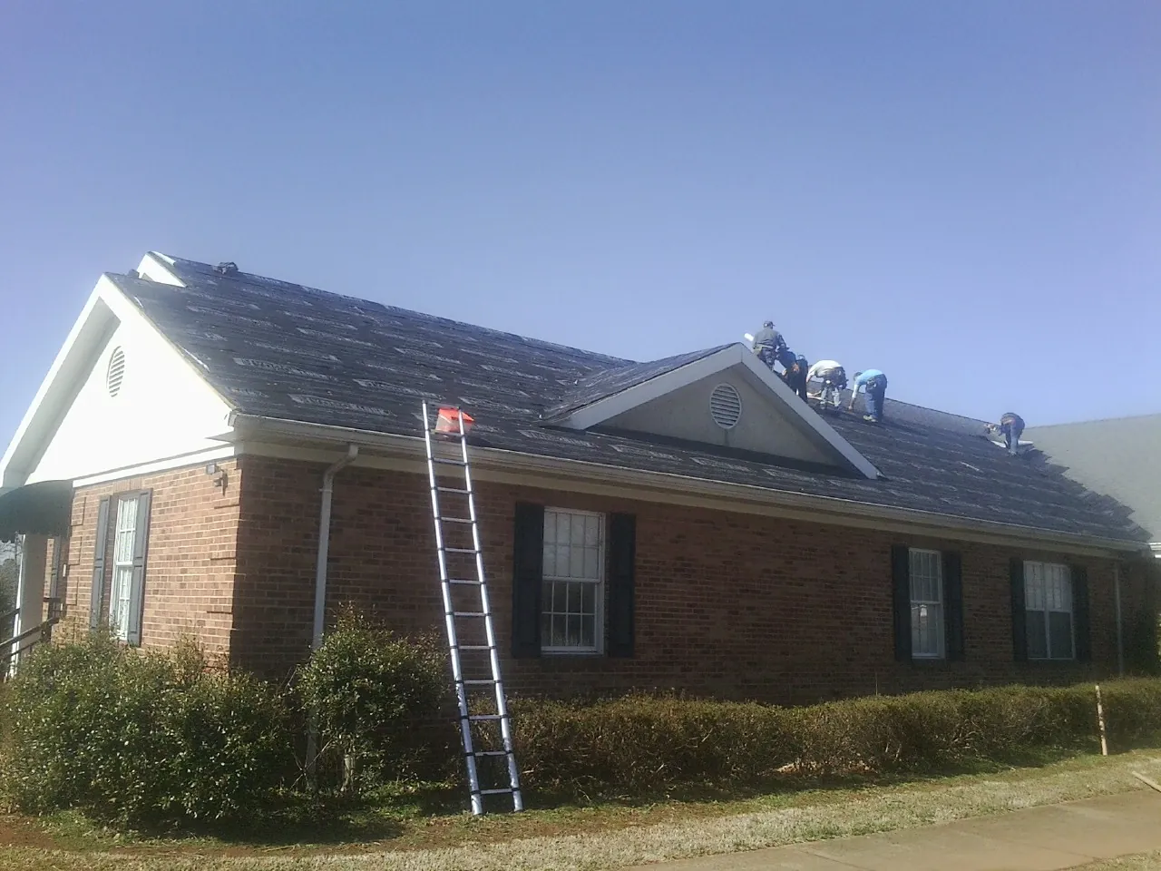 Crew tearing shingles off a brick ranch roof with a ladder set — Emergency Roof Repair work by our crew in Churubusco
