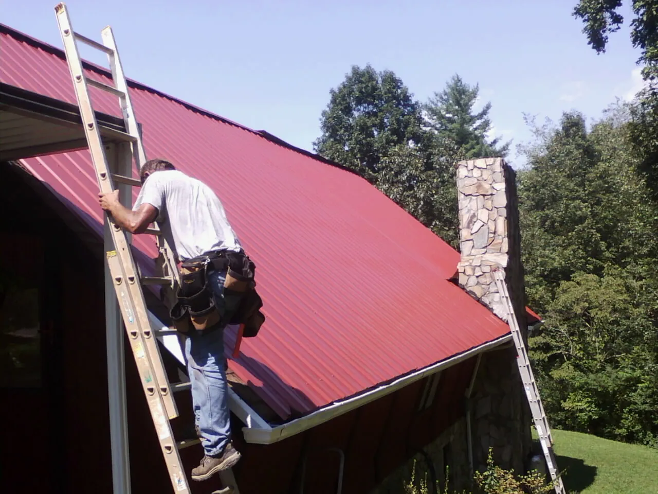 Roofer on a ladder beside a red metal roof and stone chimney — Soffit & Fascia Repair by Roofing Brothers in Churubusco